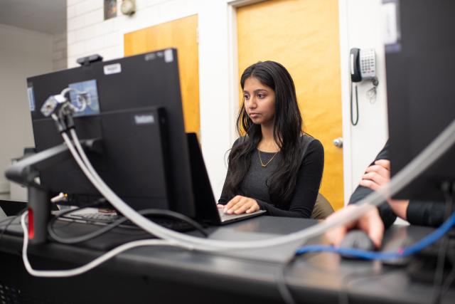 Data science student with long dark hair working in front of a desktop computer in classroom.