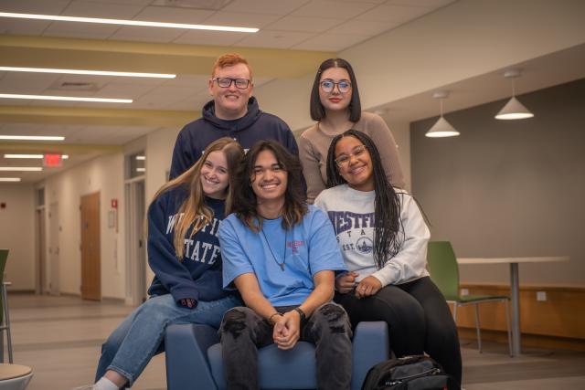 Five students wearing Westfield State sweatshirts smiling in Parenzo Hall. Two standing three sitting.