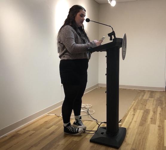 Samantha Grunden, a student reading her poetry at the Chapbook Presentation event on campus on December, 16. She is wearing a long-sleeve gray top, black pants, and converse sneakers.