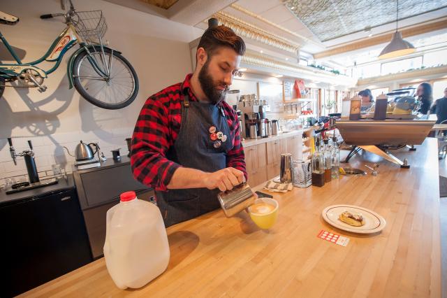 Circut Coffee Barista pouring coffee wearing black apron and black and red plaid shirt.