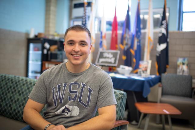 Ethan Lichwan, class of 2025. He is in the Veteran and Military office on campus, and wears a gray shirt with a blue "WSU" print on the front. He has close-cropped hair. Colored flags are blurred in the background and he is sitting on a green couch.