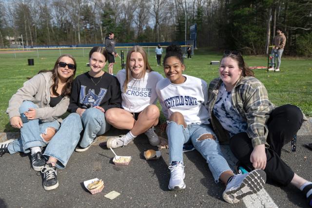 5 female students sit together and smile towards the camera during Spring Weekend activities at Westfield State University.