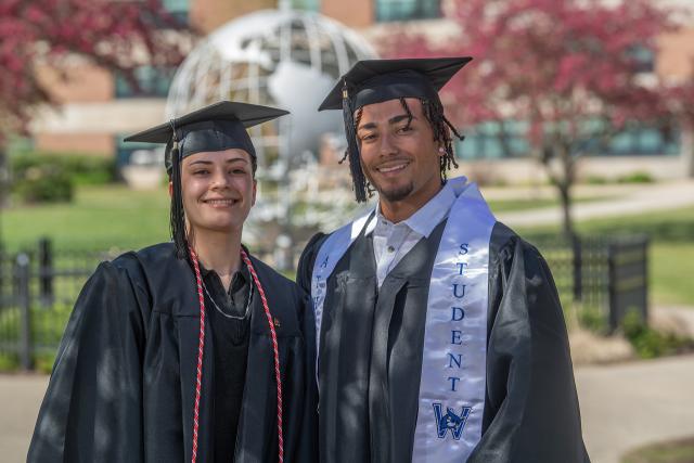 Two students smiling in front of campus globe wearing caps and gowns.