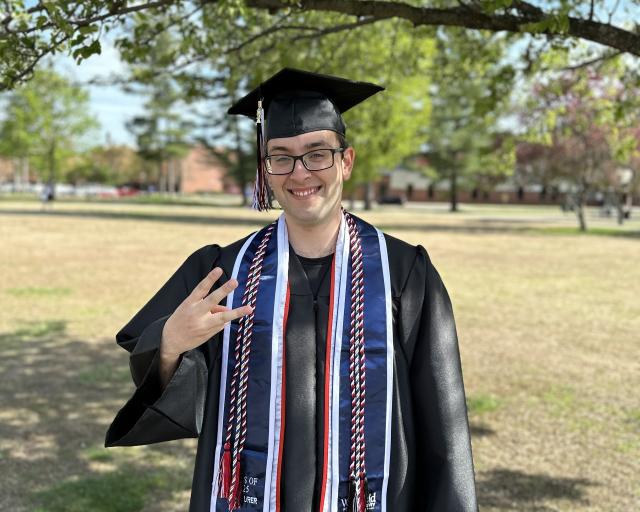 Student in cap and gown holding up three fingers to represent three days until commencement.