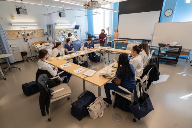 Nursing students in a classroom listening to an instructor speaking at the front of the room.