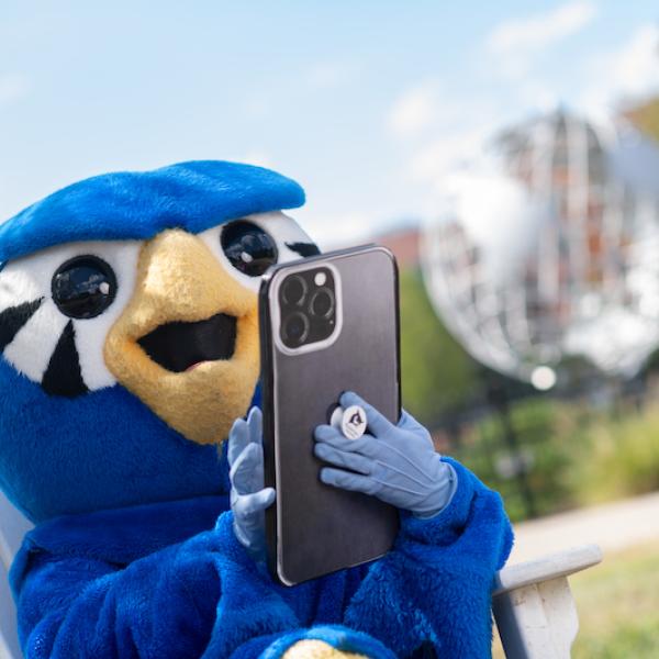 Nestor, the school mascot, wears his blue Owl suit. Nestor is holding a large phone in his hands and sits in front of the campus globe.