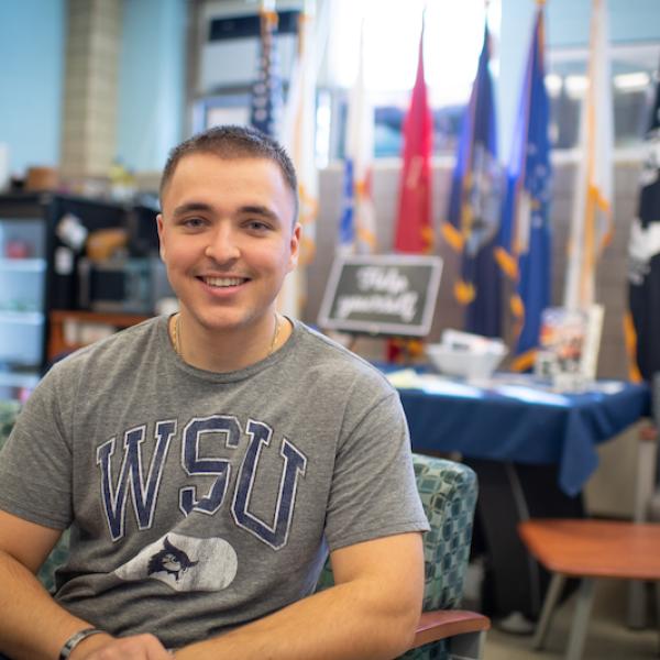 Ethan Lichwan, class of 2025. He is in the Veteran and Military office on campus, and wears a gray shirt with a blue "WSU" print on the front. He has close-cropped hair. Colored flags are blurred in the background and he is sitting on a green couch.