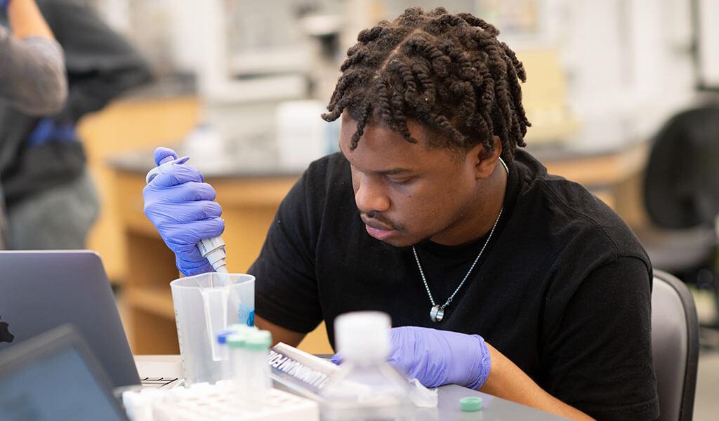 A student wearing rubber gloves conducts an experiment in a laboratory setting.