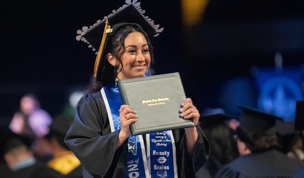 Female graduate, in mortarboard and robes, smiles as she holds up her diploma during commencement ceremonies.