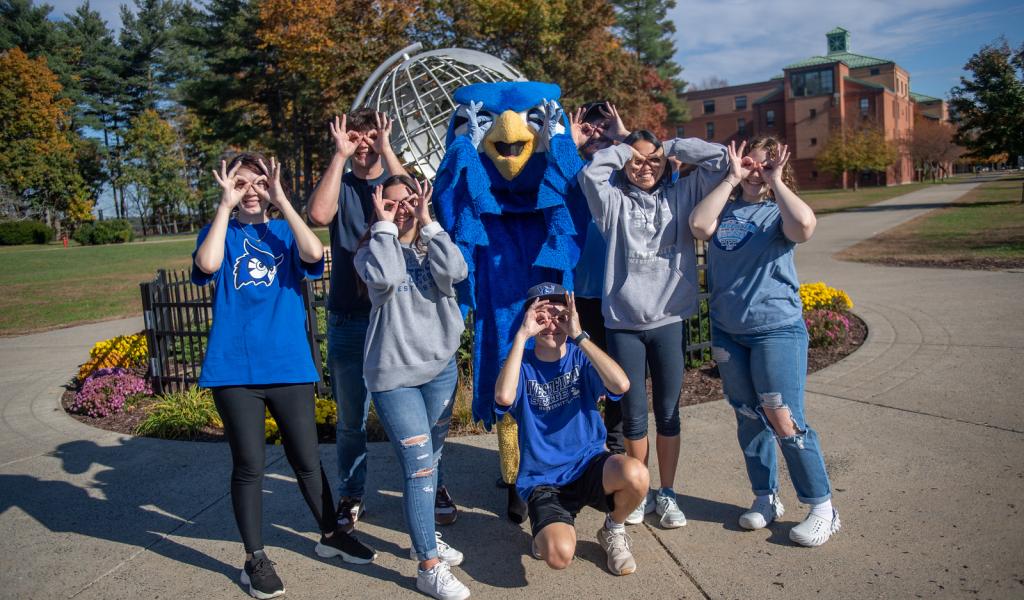 Students on the campus green with Nestor in front of campus globe using their fingers to make owl eyes.