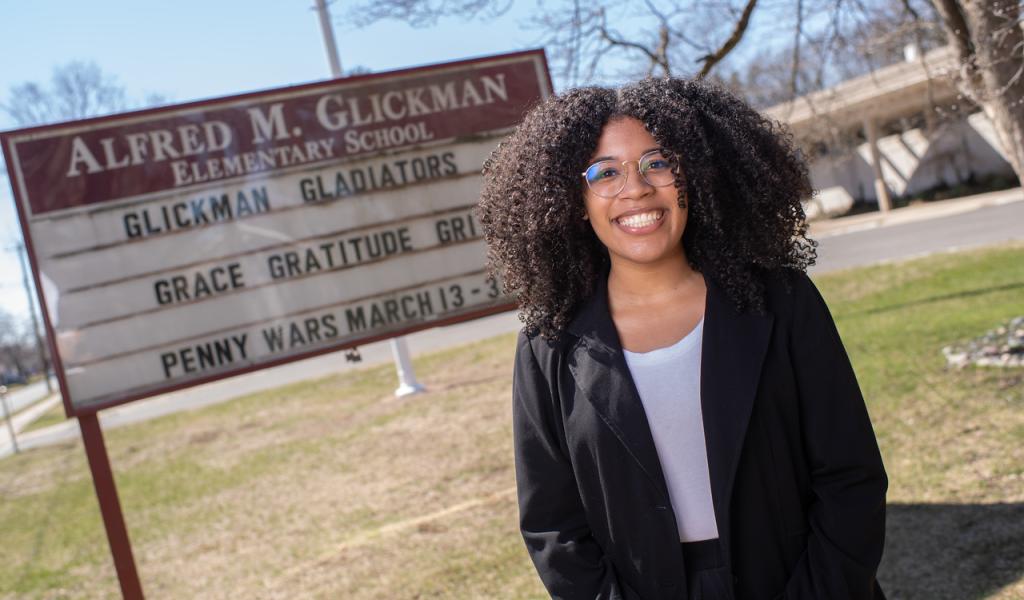 Reach to Teach alumna Lisha standing outside school smiling wearing a black coat, white shirt, and glasses.