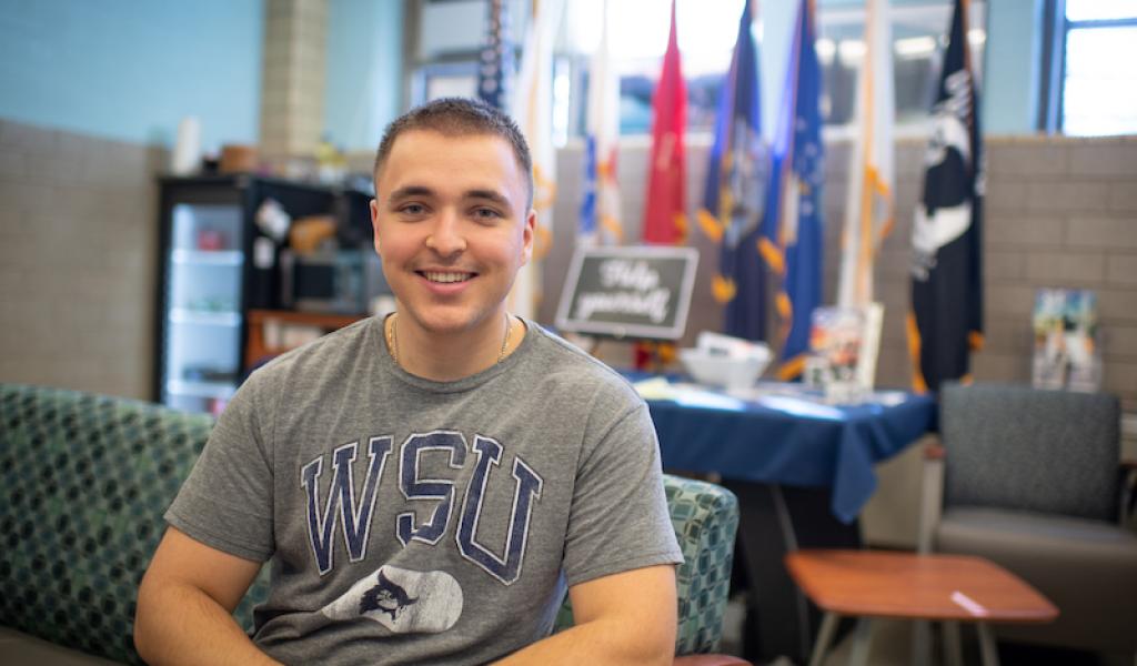 Ethan Lichwan, class of 2025. He is in the Veteran and Military office on campus, and wears a gray shirt with a blue "WSU" print on the front. He has close-cropped hair. Colored flags are blurred in the background and he is sitting on a green couch.