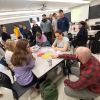 Students gather around a white table with pink and yellow pieces of paper on it. Dr. Moore, in a red and black shirt, crouches next to them.