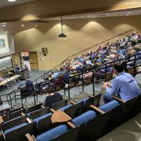 A photo of the Savignano Auditorium in Wilson Hall, with people sitting in the blue-cushioned seats. They watch a panel of guest-speakers discuss clean energy in front of them.