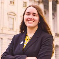 Erin Crosby wears a black, long-sleeved jacket and has her arms folded across her chest. She stands in front of a white government building, as she was featured by the US Department of Education for her work in disability services.