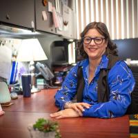 Estelle Camacho of Student Accounts. She is dressed in a long-sleeve blue shirt, blue glasses, and sits at her desk.