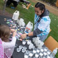 A student in a tie-die sweater makes arts and crafts outside with a young girl in a butterfly costume at a table.