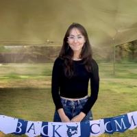 Samantha Palmer '24, in a long, black-sleeve shirt and jeans. She wears glasses and stands before a blue and white banner that says, "Welcome Back Comm Majors."