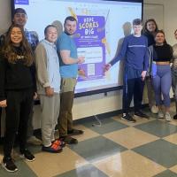 Westfield State University class of Advanced Public Relations pose in front of a poster design for “Stick Together with Rick’s Place Where Hope Scores Big,” campaign. 