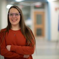 Chelsea Baker, class of 2015, leans against a white doorway. She is wearing an orange long-sleeved shirt and glasses. She's smiling directly at the camera while an out-of-focus hallway is in the background.