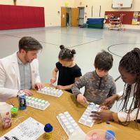 Two students in white lab coats present to the kids at Paper Elementary School in Westfield. The students and two children sit at a wooden desk and play with ice-cube trays as part of a dental hygiene demonstration.