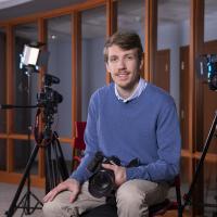 Nathan Dion, class of 2023, sits among a three professional cameras that he uses at John Garvey Inc., as their new Digital Public Relations Analyst. He wears a long-sleeved, blue sweater and tan pants. He holds a black camera in his lap while posing on a stool.