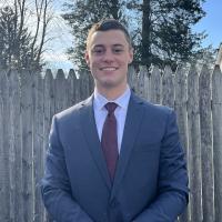 Bennett Kelly, a criminal justice major, is dressed in a blue shirt and smiles at the camera. He is outside, and a brown, wooden fence is behind him.