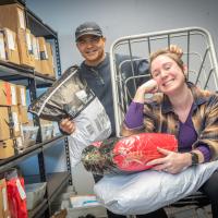 Mike DeJesus and Natalie Lussier of Mail Services, sitting on a package cart and smiling at the camera. They are surrounded by stacks of packages around them, displaying the amount of parcels needing distribution across campus.