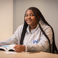 Jakayla Leary, a first-year at the University, sits at a desk with a book open before her. Her long brown hair is down, and she wears a grey sweatshirt with a blue Nestor logo on the front.