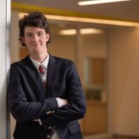 Seamus Mitchell, class of 2024 and a double-major in criminal justice and English, is dressed in a black suit coat and red tie. He is smiling and leaning against a white wall, with a golden interior behind him. Both arms are folded across his chest.