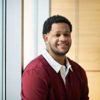 Carlos Lantigua Bari, class of 2025, is dressed in a red, long-sleeved shirt with a white collar. He's sitting near a window with wood paneling behind him. He's smiling at the camera.