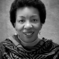 Dr. Shirley Whitaker. The photo is black and white and depicts a black woman smiling against a plain background. She wears a patterned scarf, dangly earrings, and is smiling at the camera.