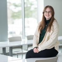 Nicole Dolat, class of 2024. She is dressed in a green shirt, a white over-sweater, black glasses, black pants, and sits on top of a white, glossy desk. The walls behind her are white, and she is in a classroom. Windows along it are open, and there is greenery blurred in the background.