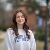 Paige Freeman, a senior at the University. She is outside, near the Ely building. She has long, brown hair and a gray sweatshirt which says "Westfield State" in blue lettering. Blurred out trees, grass, and the sidewalk are in the background.