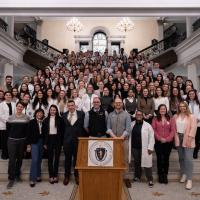 2024 Lobby Day, an event for Physician Assistants and students studying to be PAs. A large group of people, some in white coats, gather on the steps of the venue in Boston, MA. A wooden podium sits before all of them. Two chandeliers hang in the background behind them.