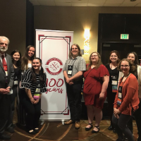 The 2023 Sigma Tau Delta Centennial Convention. Dr. Glenn Brewster, along with Alexis Crafts, Sam Grunden, Chloe Sanfacon, Victoria Nesmelova, and Julia Robak. They stand, pose, and smile in front of a small white banner which says, "100 years" in red script. 