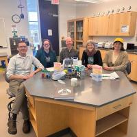 From left to right: Dr. Arne Christensen, Dr. Megan Kennedy, Dr. Volker Ecke, Dr. Christine Von Renesse, and Dr. Shannon Gleason. They sit in a Biology lab, surrounded by tan desks and cabinets along the walls. They sit together at one desk, wearing blue lanyards and smiling at the camera as part of an interview on the University's new STEM-ACT grant.