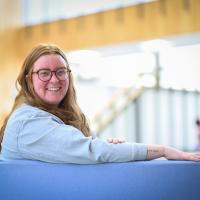 Averie Bye-Dickinson, a graduate student of the Master go Public Administration program. She is sitting in the lobby in Horace Mann, and is wearing a gray, long-sleeve shirt. She has red hair and glasses, and has one arm over the back of the chair she sits on as she smiles at the camera. White light from the background streams in behind her.