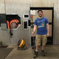 Brian Mernoff, an alum of the University. He is standing next to heavy machinery and is wearing a blue T-shirt and tan cargo shorts. He's smiling while holding a staff.