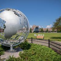 A stock photo of the silver campus globe. The sky above is blue, with green grass and vegetation around he globe itself, wrapped in by a black fence. Ely is in the distance, with a tree to the side of the frame.