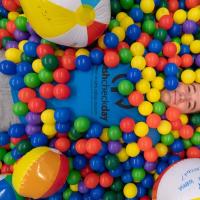 A male student from the 2023 Fresh Check Day. He is wearing a dark blue shirt with black lettering that says, "Fresh Check Day", and is laying in a colorful ball pit while smiling at the camera.