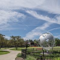 The campus globe amidst a sunny, summer day. The sky is blue and cloudy, and trees and grass dot the campus around it.