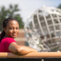 Marilyn Maison, class of 2024. She is sitting outside, on a bench in front of the silver, campus globe. The background behind her is blurred, and she has one arm slunk over the railing she leans against.