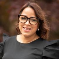 Assistant Director for Student Access and Support, Marjorie Rodriguez. She wears a black shirt and glasses and stands in front of blurred foliage.